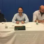 Three men sitting at a table with a sign