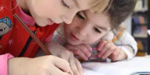 Two children are writing on a piece of paper.