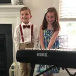 Two children standing next to a piano keyboard.