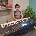 A young boy is standing next to an electronic keyboard.