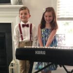 Two children standing next to a piano keyboard.